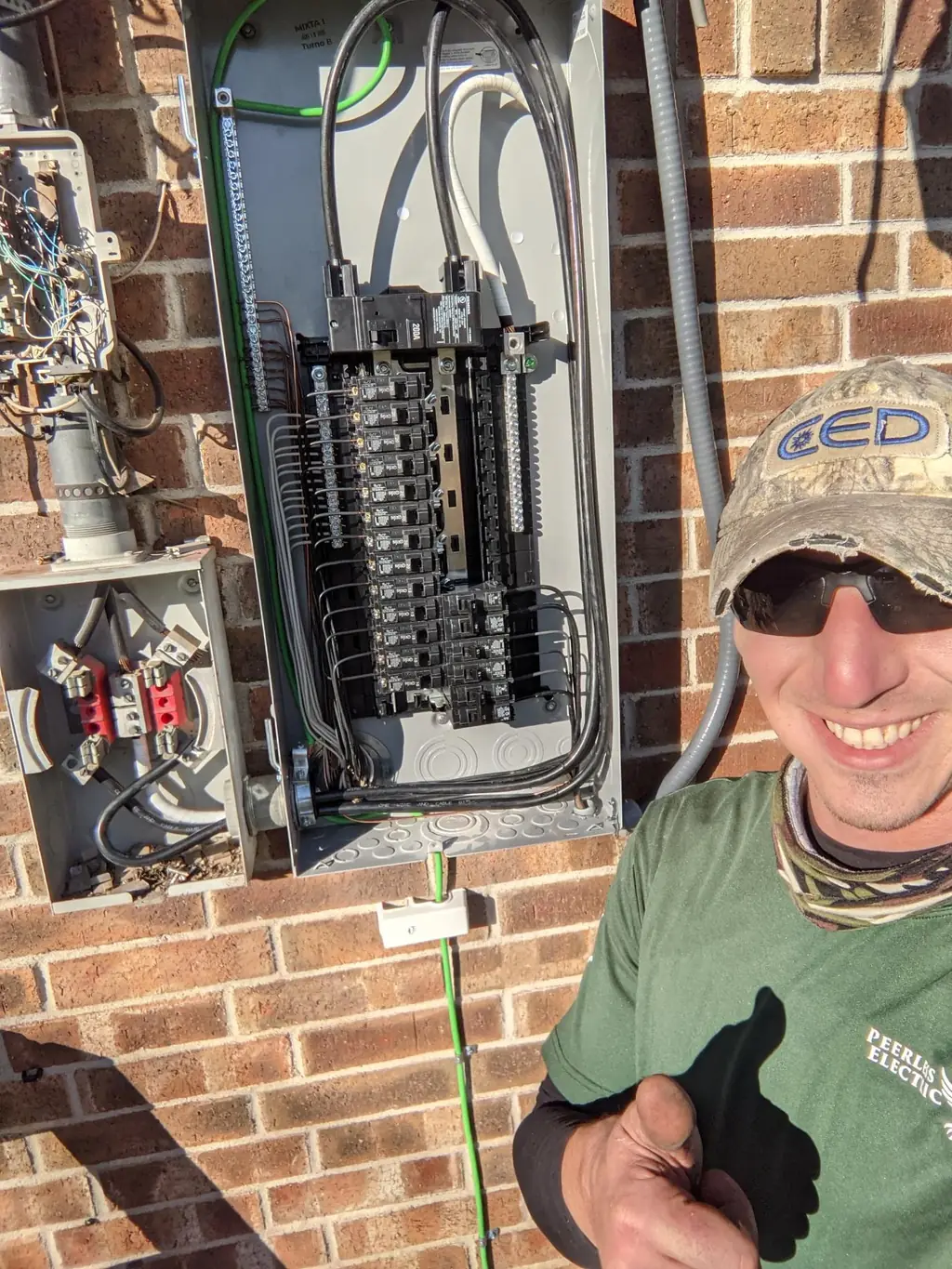 Peerless Electric technician beside a finished 200 amp exterior panel install on a brick wall